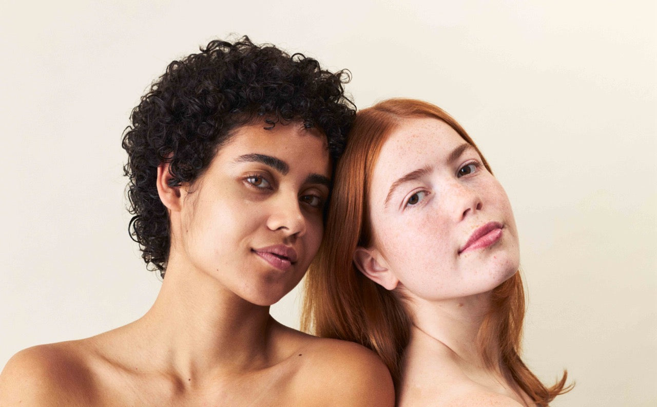 Zwei Frauen, die aneinander lehnen und in die Kamera schauen, die eine hat einen normalen Hauttyp und die andere hat Mischhaut.