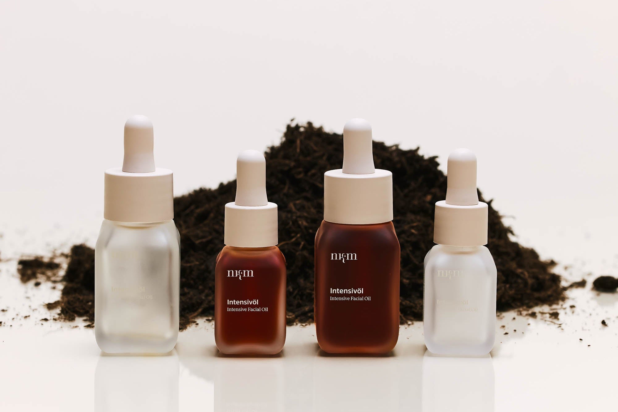Im Hintergrund Erde, im Vordergrund das Intensivöl in groß und in Reisegröße. Links und rechts davon stehen leere Glasflaschen