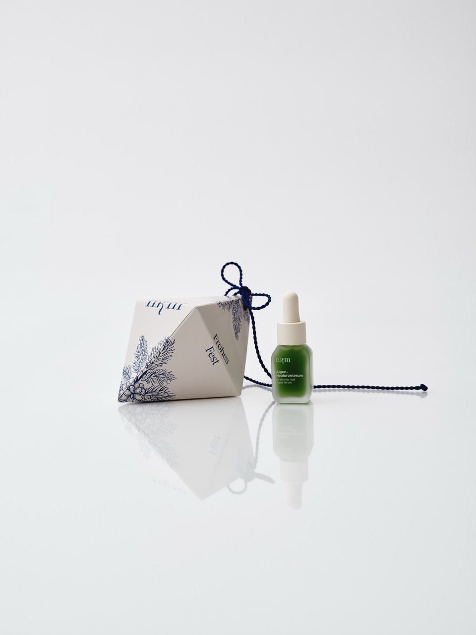 Weißes Weihnachtsornament mit aufgemalten blauen Tannenzweigen, daneben das Algen-Hyaluronserum von nkm