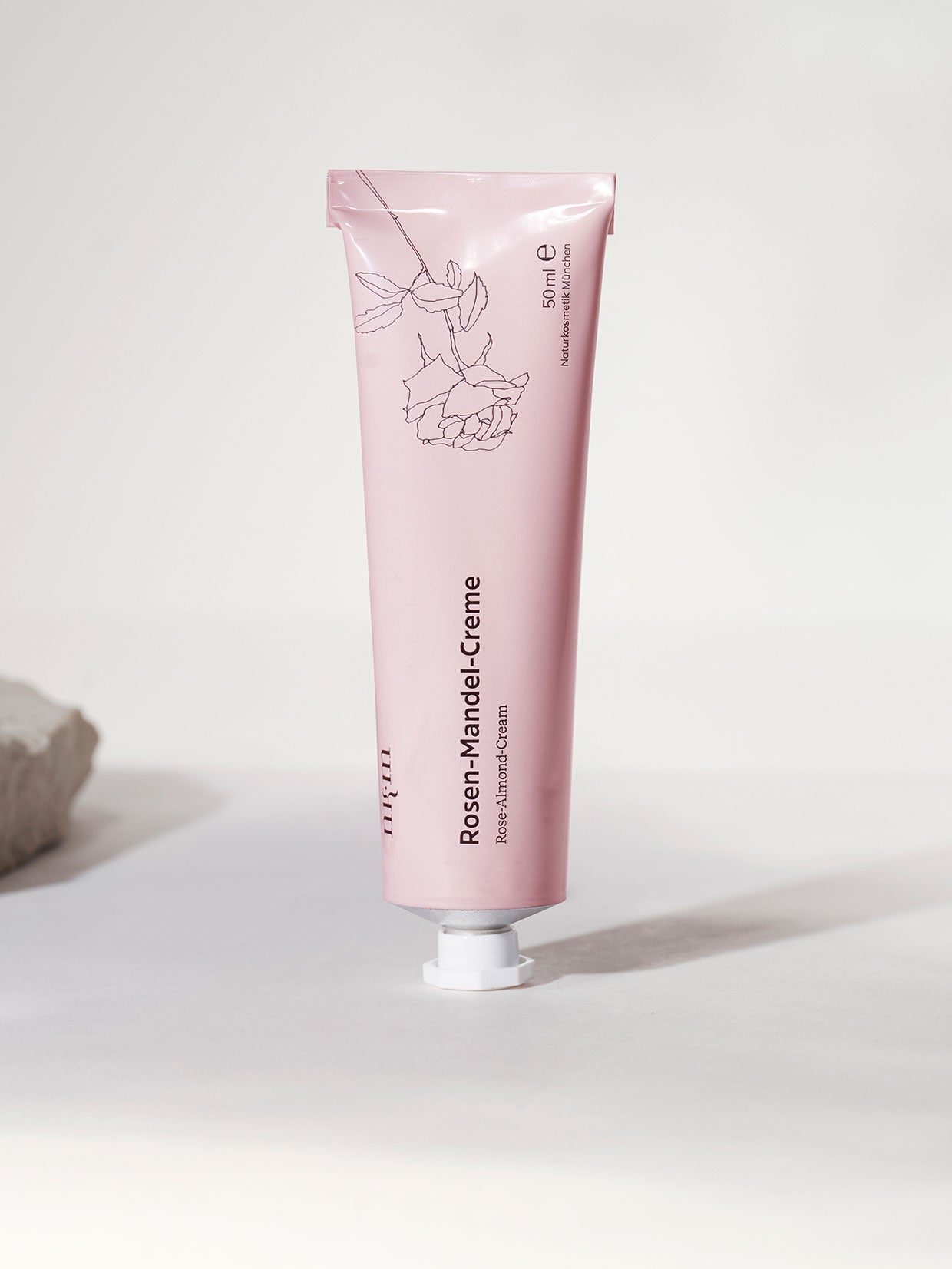 Eine rosafarbene Tube Rosen-Mandel-Creme der nkm Naturkosmetik München GmbH steht aufrecht auf einer hellen Fläche und spendet Feuchtigkeit in minimalem Rahmen.