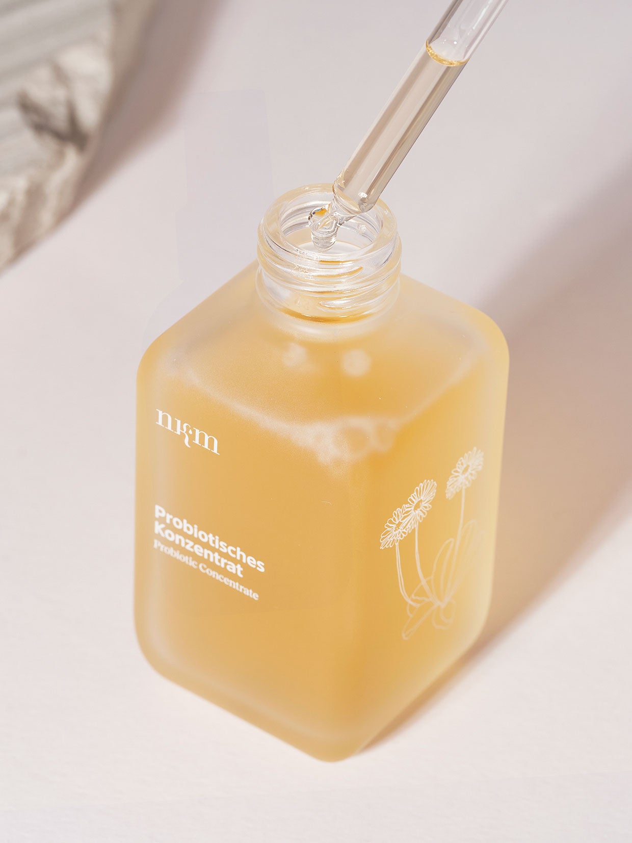 Auf einer hellen Fläche steht ein Flakon von nkm Naturkosmetik München GmbH Probiotisches Konzentrat mit floralem Design und klarem Tropfer, perfekt für die Hautbarriere.