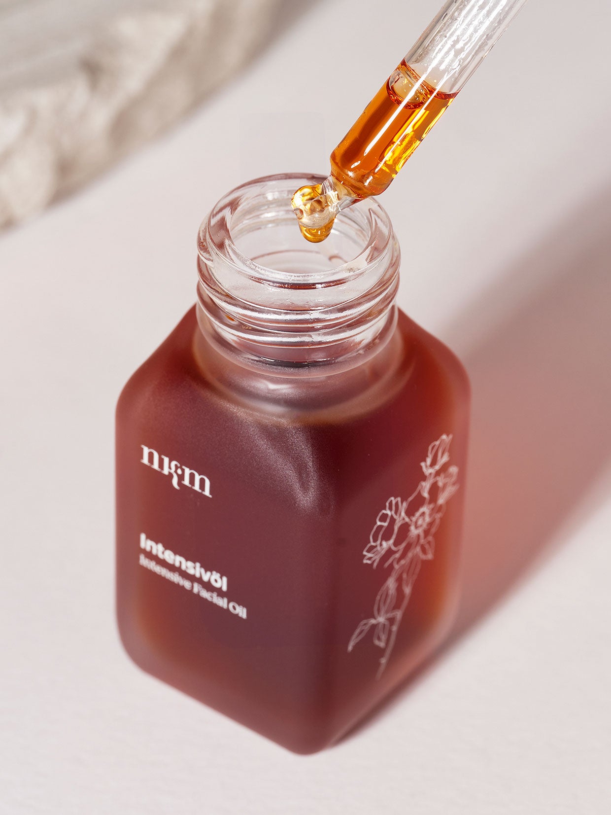 Ein Tropfer spendet das Öl in eine mattbraune Flasche mit der Aufschrift Reisegröße Intensivöl von nkm Naturkosmetik München GmbH, die mit einem floralen Design verziert ist und pflegendes Sanddorn- und Rosenöl enthält.