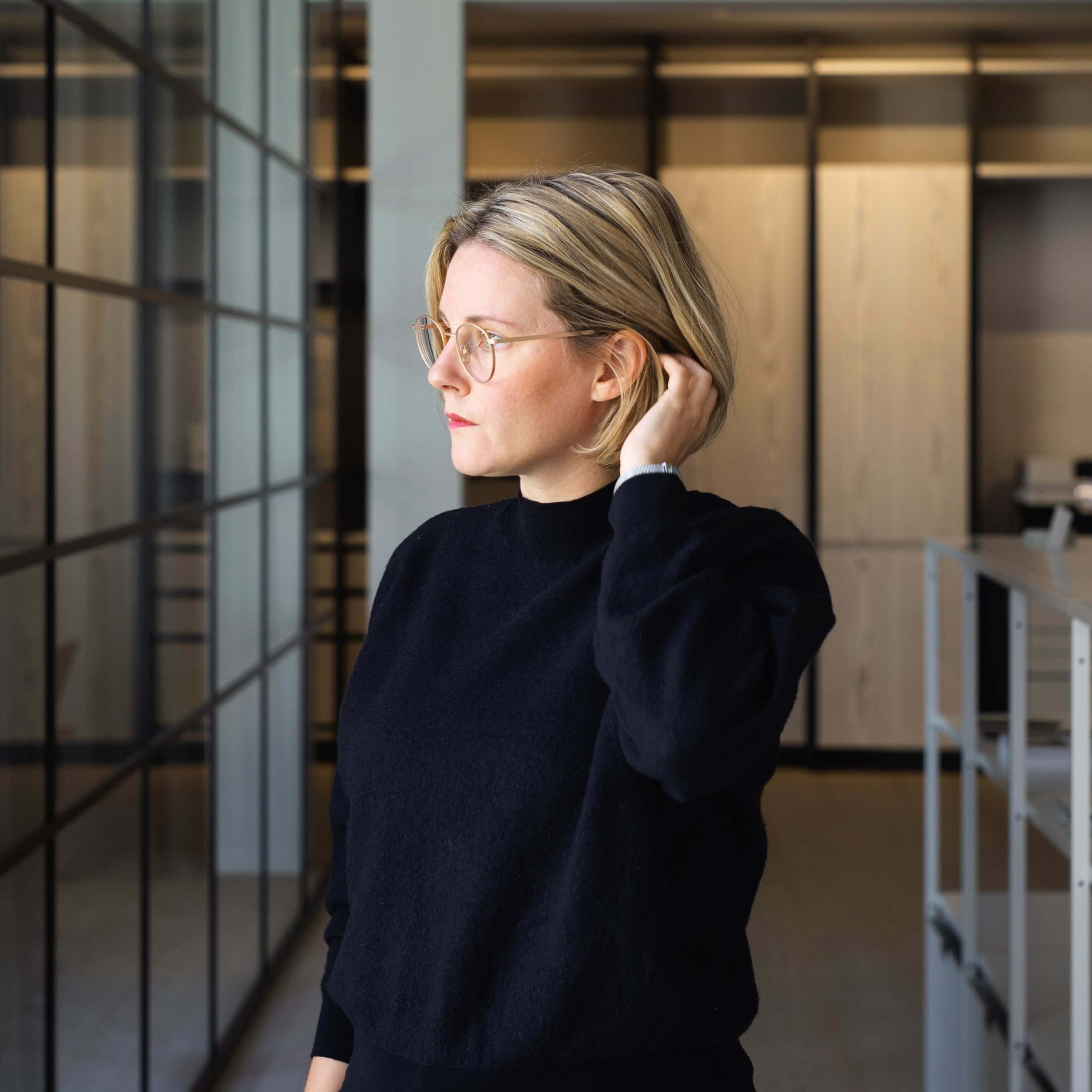 Frau mit kurzen blonden Haaren und Brille steht vor einem Fenster
