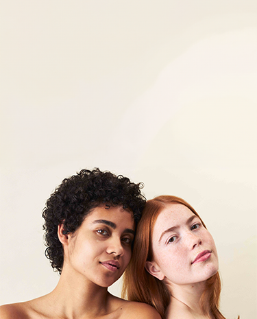 Zwei Frauen, die aneinander lehnen und in die Kamera schauen, die eine hat einen normalen Hauttyp und die andere hat Mischhaut.