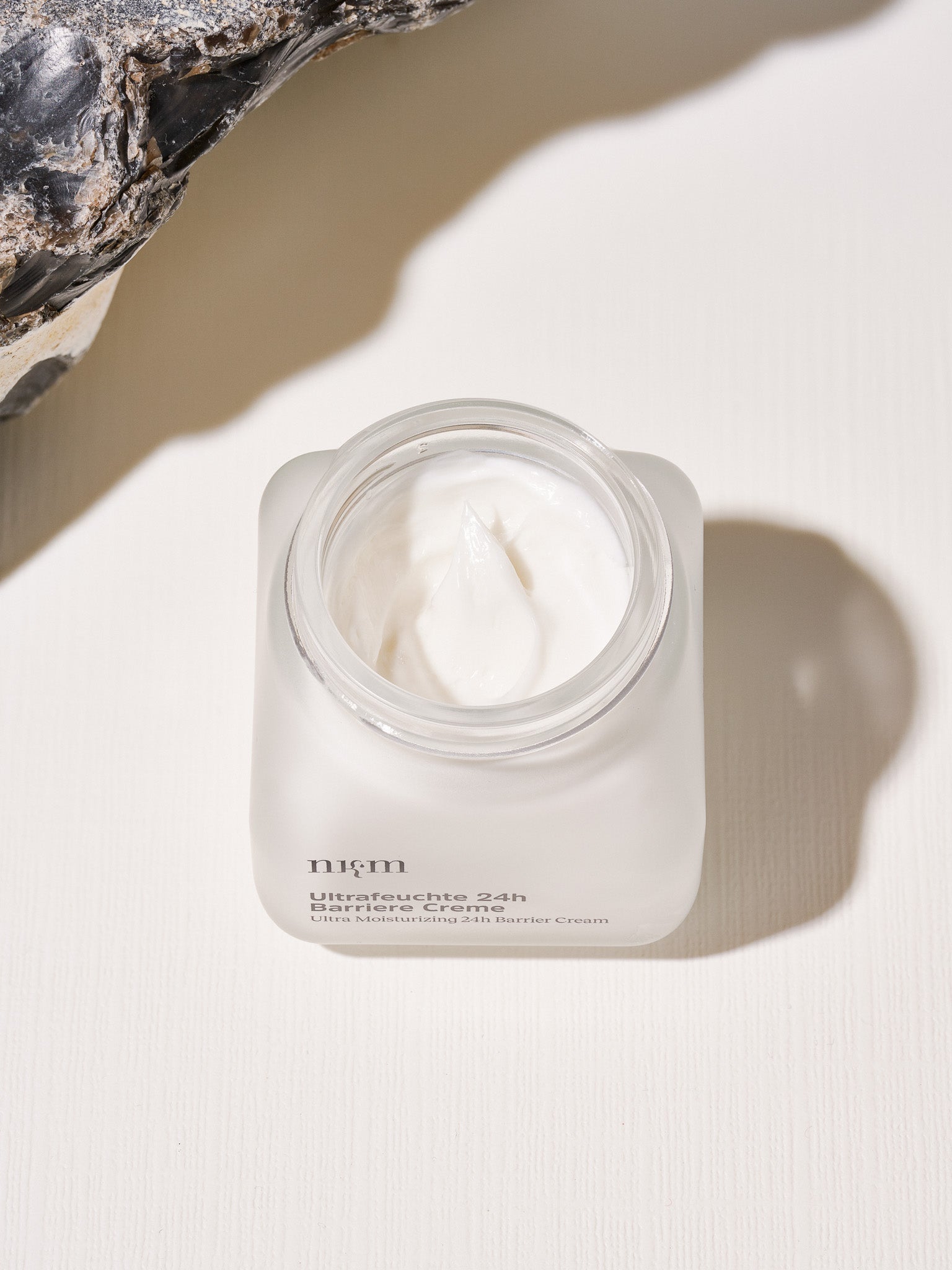 Ein geöffneter Glastiegel der nkm Naturkosmetik München GmbH Ultrafeuchte 24h Barriere Creme steht auf einer leicht strukturierten Fläche neben einem Stein.