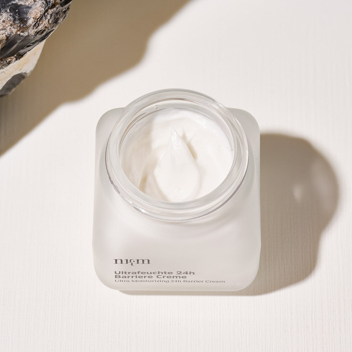 Ein geöffneter Glastiegel der nkm Naturkosmetik München GmbH Ultrafeuchte 24h Barriere Creme steht auf einer leicht strukturierten Fläche neben einem Stein.