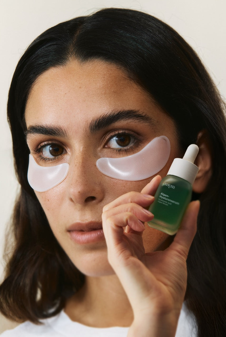 Eine Frau, die das Algen-Hyaluronserum in der Hand hält und die Augenpads unter den Augen aufgetragen hat.