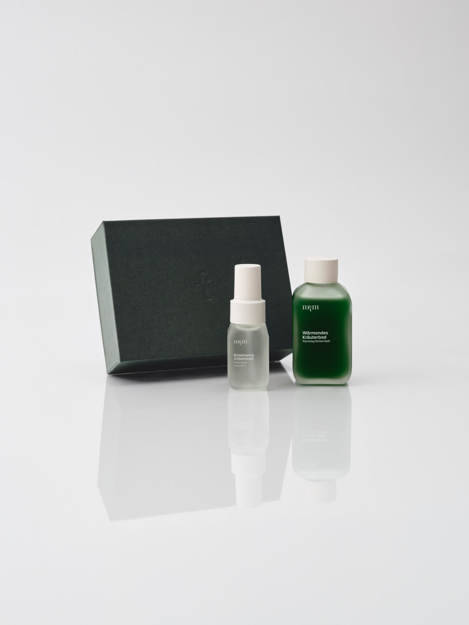 Eine dunkelgrüne Box und davor das Wärmende Kräuterbad und Kissenspray Zirbenwald von nkm