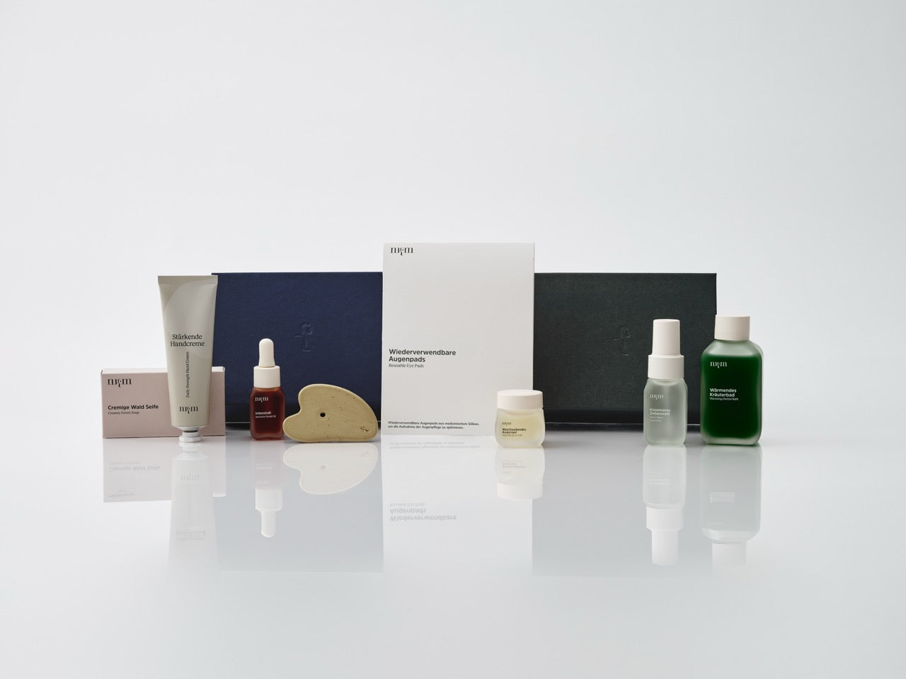 Eine dunkelblaue und eine dunkelgrüne Box. Davor angeordnet: Cremige Wald Seife, Stärkende Handcreme, Wiederverwendbare Augenpads, Intensivöl, Abschwellendes Augengel, Kissenspray Zirbenwald, Wärmendes Kräuterbad und Gua Sha