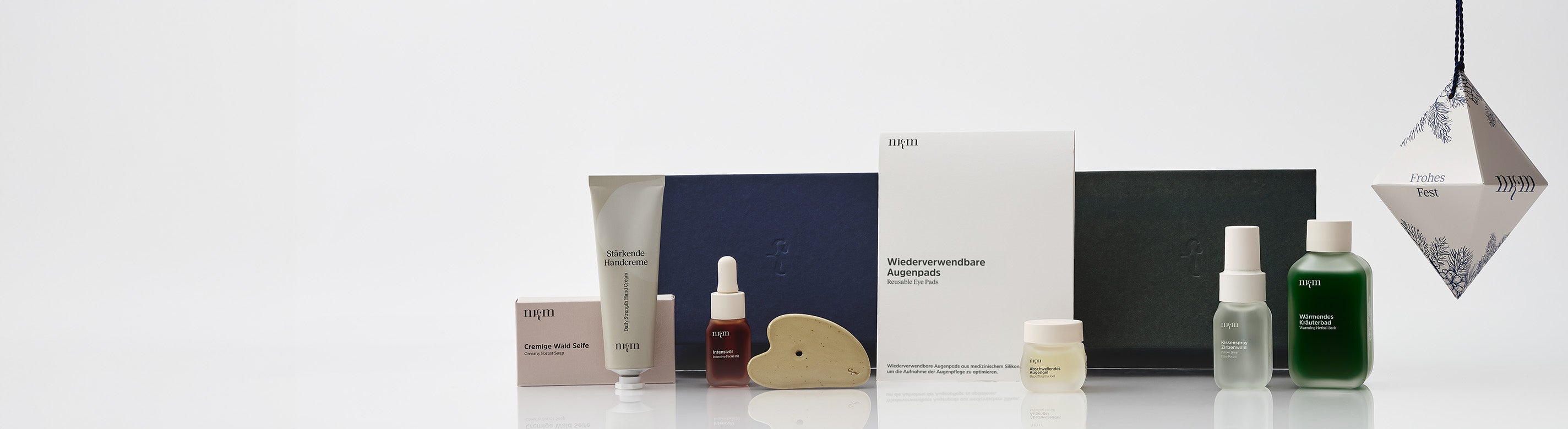 Blaue und grüne Weihnachtsbox, davor Cremige Wald Seife, Stärkende Handcreme, Intensivöl, Gua Sha, Wiederverwendbare Augenpads, Abschwellendes Augengel, Kissenspray Zirbenwald und Wärmendes Kräuterbad. Rechts das weihnachtliche Ornament.
